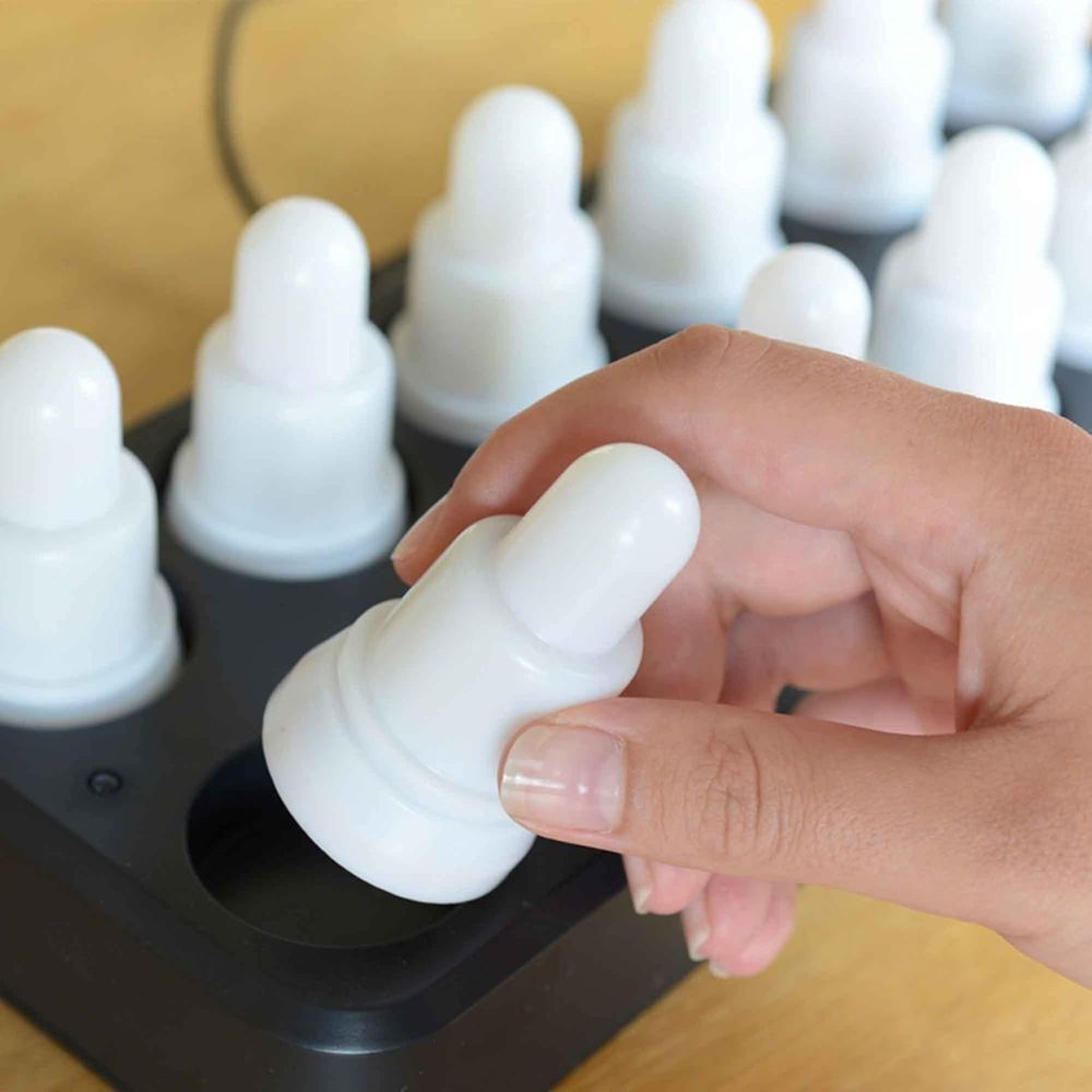 Een hand plaatst of verwijdert een witte, ronde LED-kaars uit het zwarte dienblad van de Duni GmbH LED-set, warmwit & multicolor – 12 stuks kleurveranderende lampjes; elke identieke, oplaadbare kaars staat op een houten voetstuk.