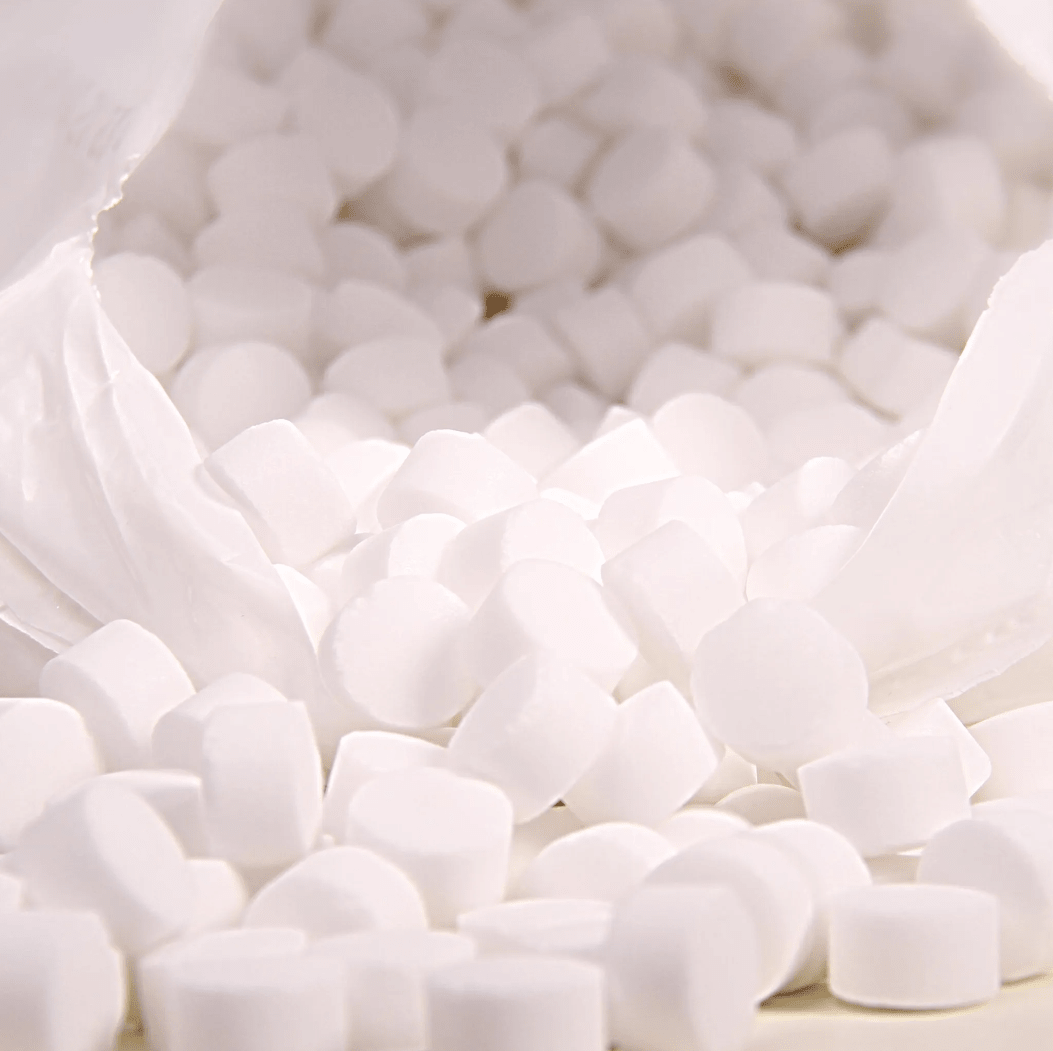 Een close-up toont een witte plastic zak met het opschrift „K+S Minerals and Agriculture GmbH“, die Regenit-kookzouttabletten volgens EN 973 type | zak (25 kg) bevat, die op een oppervlak vallen. De kleine, ronde tabletten hebben een uniforme vorm en grootte en zijn ideaal voor gebruik in wateronthardingsinstallaties.