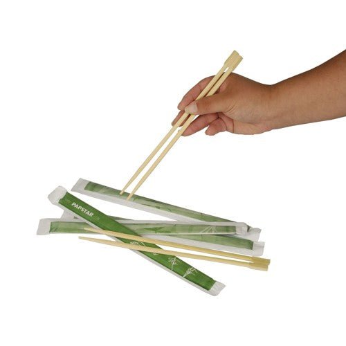 Een hand houdt houten PAPSTAR eetstokjes "pure" 23 cm boven meerdere paren, elk afzonderlijk verpakt in groene papieren hoezen. Product: PAPSTAR eetstokjes "pure", 50 paar | Folie van PAPSTAR GmbH, afgebeeld op een witte achtergrond.