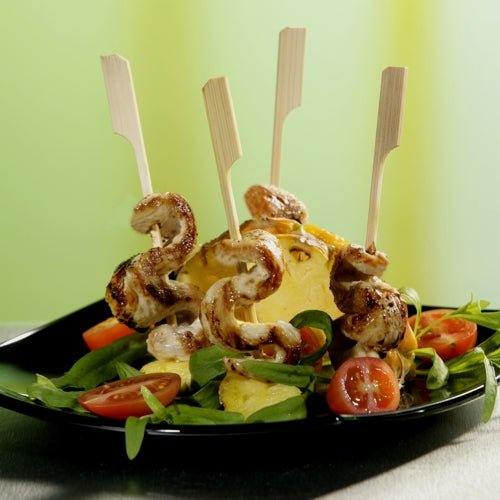 Een zwart bord met salade, gehalveerde kerstomaten, ananas en PAPSTAR GmbH fingerfood-spiesen "puur" 10 cm (40 stuks) - gegrilde kippenspiesen verticaal gerangschikt. Ideaal voor golfevenementen en catering. De groene achtergrond is wazig.