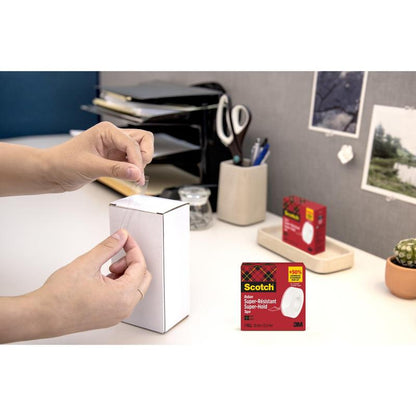 Een persoon sluit een witte doos met kantoormaterialen met Scotch® Super-Hold Secure Tape, 1 rol, 19 mm x 25,4 m van 3M Deutschland GmbH. De verpakking van de plakband is in de rechter benedenhoek ingevoegd.