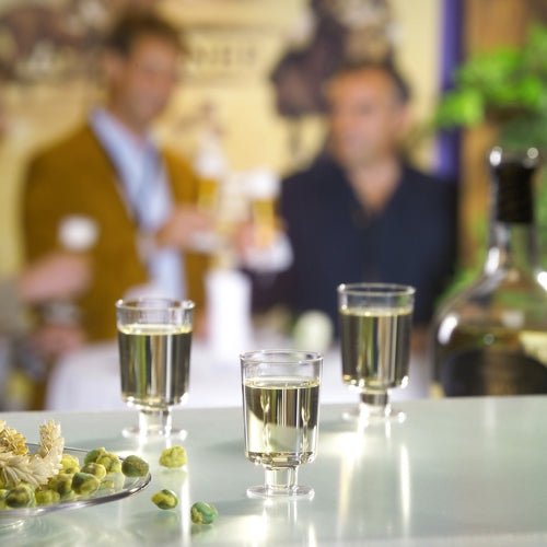 De Starpak 20 steelglazen voor witte wijn van glashelder polystyreen staan op een tafel met olijven en een fles, terwijl drie personen op de achtergrond met drankjes praten.