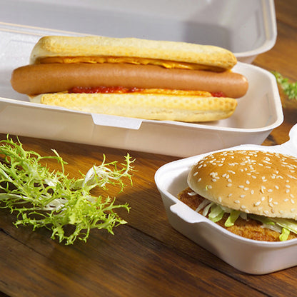 Een hotdog met ketchup en mosterd wordt geserveerd in een Starpak 100 Allzweckbox met klapdeksel XPS 1800 ml, naast een sesam-hamburger met sla in een andere Starpak-box. Op de houten tafel ligt een beetje gekruld slablad.