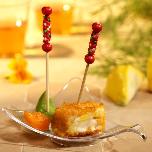 Een heldere glazen schaal toont een gepaneerd voedingsblokje en twee stukjes fruit, elk met een PAPSTAR Fingerfood picker "Mexico" (7,5 cm) van PAPSTAR GmbH, perfect voor feestjes. In de zacht vervaagde achtergrond zijn drankjes en een citroenpartje te zien.