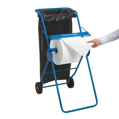 Een blauwe metalen dispenser houdt een WypAll® L10 papieren doek voor oppervlakken - grootrol (38 x 23,5 cm) van Kimberly-Clark GmbH en een zwarte vuilniszak; een persoon trekt een vel voor de oppervlaktereiniging eruit.