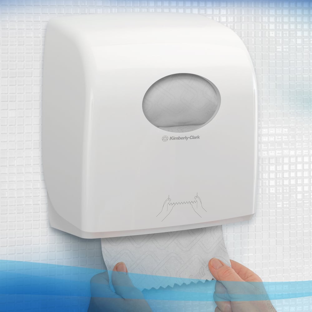 Een persoon trekt een handdoek uit een witte Kimberly-Clark GmbH Scott® Essential™ handdoekdispenser met een klein ovaal venster, waardoor je de papierrol binnenin kunt zien, met gestructureerde tegels op de achtergrond. 6 witte rollen per doos.