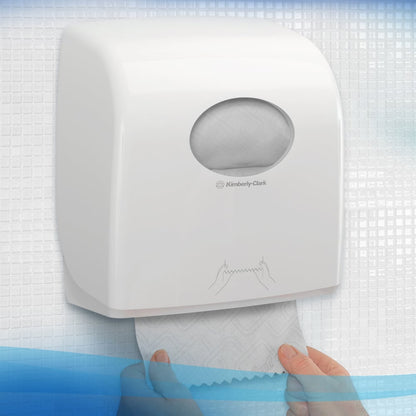 Een persoon trekt een handdoek uit een witte Kimberly-Clark GmbH Scott® Essential™ handdoekdispenser met een klein ovaal venster, waardoor je de papierrol binnenin kunt zien, met gestructureerde tegels op de achtergrond. 6 witte rollen per doos.