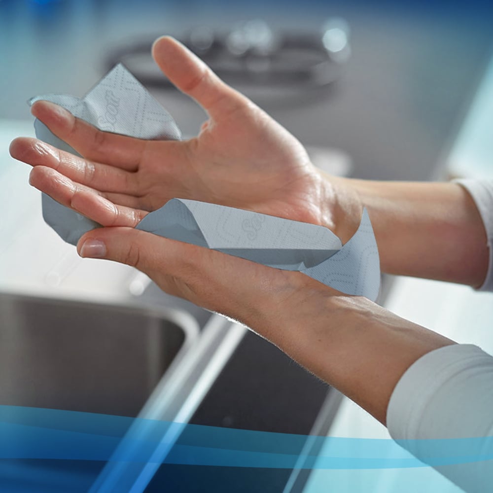 Een persoon droogt zijn handen met Scott® Essential™ handdoeken van Kimberly-Clark GmbH (rol / blauw - 6 rollen) boven een wastafel in een moderne keuken.