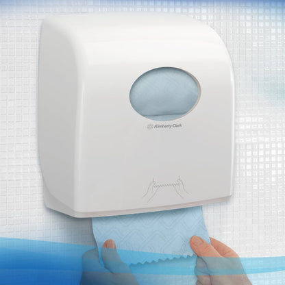 Een persoon trekt een blauwe Scott® Essential™ handdoek - rol uit een witte dispenser van Kimberly-Clark GmbH, die is bevestigd aan een betegelde muur met een rond venster dat de daarin aanwezige handdoeken en een illustratie voor handdoekuitgifte toont.