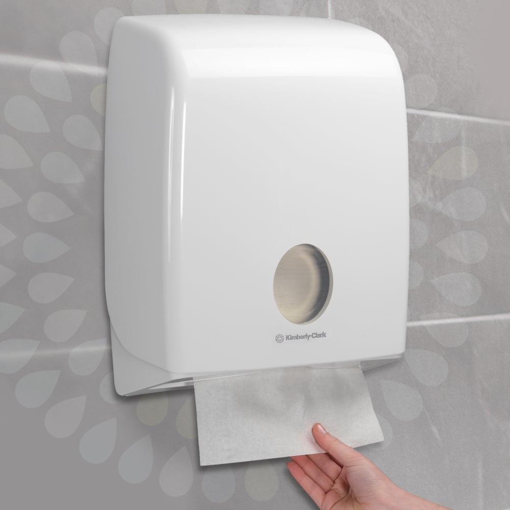Een hand trekt een Kleenex® Ultra™ Handdoek uit een aan de muur gemonteerde witte dispenser van Kimberly-Clark GmbH, die aan een betegelde muur is geïnstalleerd (Interfold/Wit/Groot - 30 clips x 94 vellen | doos, 30 pakjes).