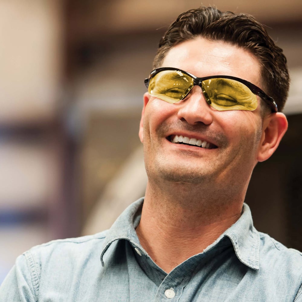 Ein Mann in einem hellblauen Hemd lächelt in einem Haus und trägt Auslaufartikel der Kimberly-Clark GmbH: KleenGuard® V30 Nemesis™ Schutzbrille - Caramel, wobei er seinen Fokus auf den Augenschutz legt. Der Hintergrund ist unscharf.