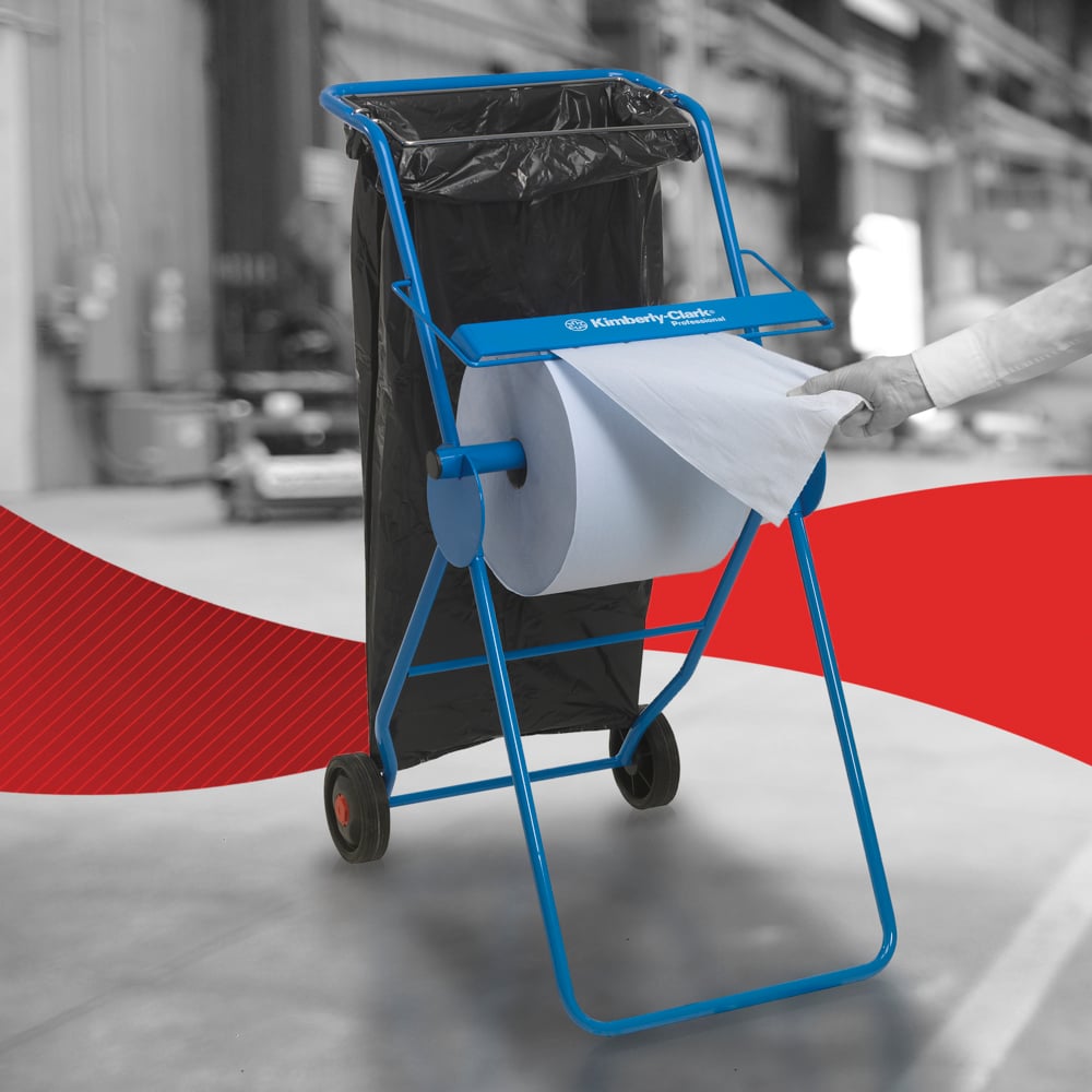 Een persoon trekt een WypAll® L10 papieren doek voor oppervlakken - grootrol (38,00 x 23,50 cm) van Kimberly-Clark GmbH van een blauwe metalen standaard met wielen en een daaraan bevestigde zwarte vuilniszak, wat wijst op een efficiënte oppervlaktereiniging in industriële omgevingen.