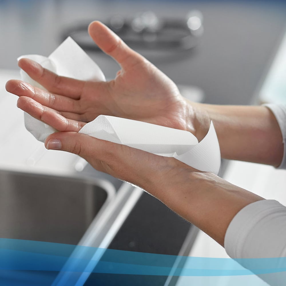 Iemand droogt zijn handen met Scott® Control™ Handdoeken - Interfold / Wit / Klein van Kimberly-Clark GmbH, met een keukentafel en een kraan op de achtergrond. De verpakking bevat 15 x 304 vellen per doos.