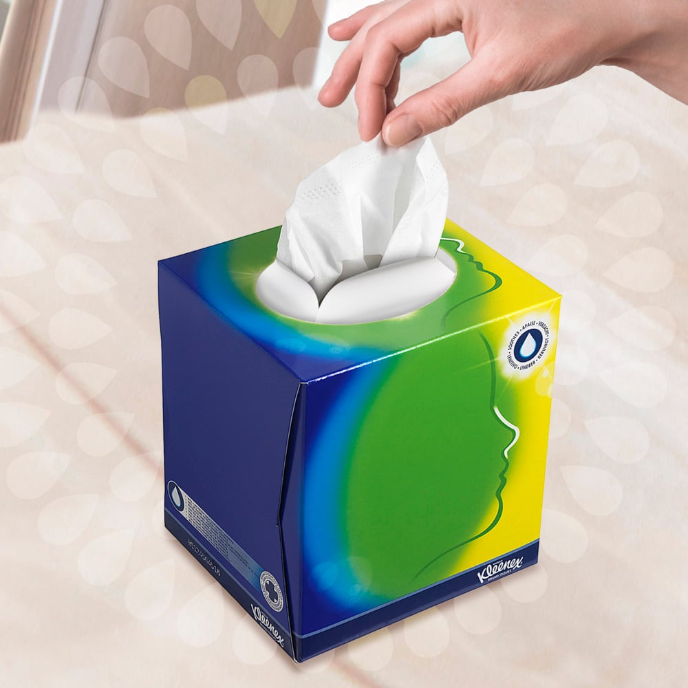 Een hand trekt een tissue uit de Kleenex® cosmetische tissues - kubusdoos / wit van Kimberly-Clark GmbH met een blauw, groen en geel ontwerp en een witte ovale opening. Een gestileerd profiel wijst op gezichtsverzorging voor een zacht vervaagde achtergrond.