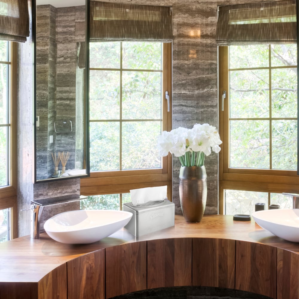 Ein modernes Badezimmer mit einer hölzernen Arbeitsplatte, zwei weißen Waschbecken, braunen Jalousien, einer Vase mit weißen Blumen und einem eleganten Kleenex® Handtuchspender aus Edelstahl von Kimberly-Clark GmbH mit Zupfbox-Funktion neben der Tissue-Box. Draußen sind Bäume zu sehen.