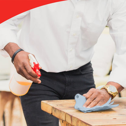 Een persoon met een wit shirt en horloge reinigt een houten tafel met een blauwe doek, mogelijk WypAll® L20 General Clean - BRAG™ Box van Kimberly-Clark GmbH. De afbeelding heeft bovenaan een rode gebogen rand die de aandacht voor detail benadrukt.