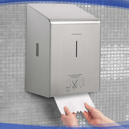 Een persoon trekt papieren handdoeken uit een Kimberly-Clark Professional™ rollenhanddoekdispenser - roestvrij staal van Kimberly-Clark GmbH, die te midden van kleine grijze vierkante tegels aan de muur is bevestigd.