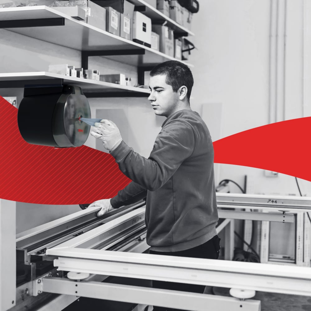Een man bedient machines in een werkplaats. Op de achtergrond zijn planken met gereedschap en het WypAll® L20 General Clean Centrefeed for Reach+™ (blauw, 2-laags, 6x550 vel) van Kimberly-Clark GmbH te zien te midden van rode grafische elementen.