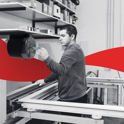 Een man bedient machines in een werkplaats. Op de achtergrond zijn planken met gereedschap en het WypAll® L20 General Clean Centrefeed for Reach+™ (blauw, 2-laags, 6x550 vel) van Kimberly-Clark GmbH te zien te midden van rode grafische elementen.