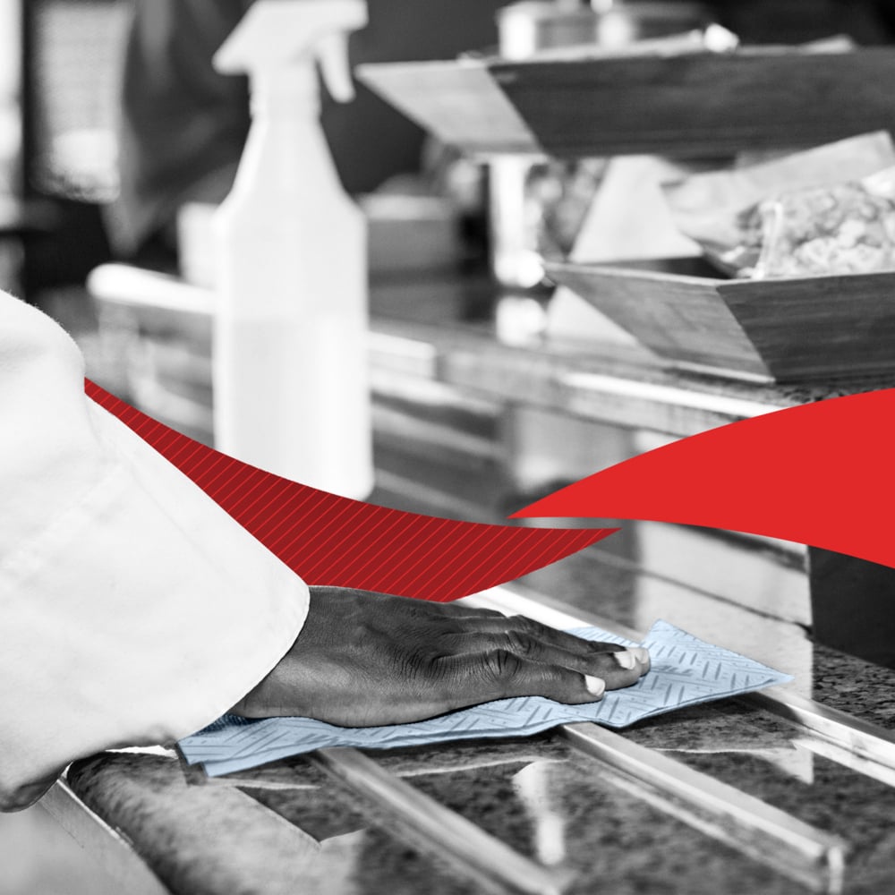 Een persoon veegt een toonbank af met een blauwe doek en toont reiniging. Op de toonbank bevinden zich een WypAll® L20 General Clean - Centrefeed for Reach+™ rol en trays van Kimberly-Clark GmbH met een rood grafisch element in het midden van de afbeelding.