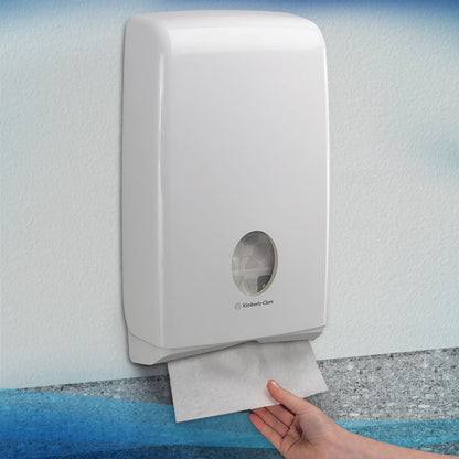 Een hand trekt een Scott® SLIMFOLD-handdoek uit een witte Kimberly-Clark GmbH wanddispenser met kijkvenster, die gevouwen doeken aan de binnenkant toont. De hygiënische dispenser is aan een lichte muur gemonteerd; hij bevat 16 clips x 110 vellen per karton.