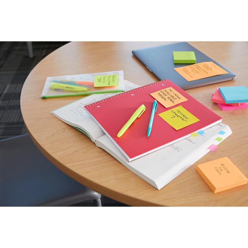 Een ronde houten tafel met een rood notitieboek, een geopend leerboek, pennen, markeerstiften, een laptop, een transparante map en 3M Deutschland GmbH Post-it® Super Sticky Z-Notes (Boost Collection, 76x76mm) is te zien; een stoel is gedeeltelijk afgebeeld.
