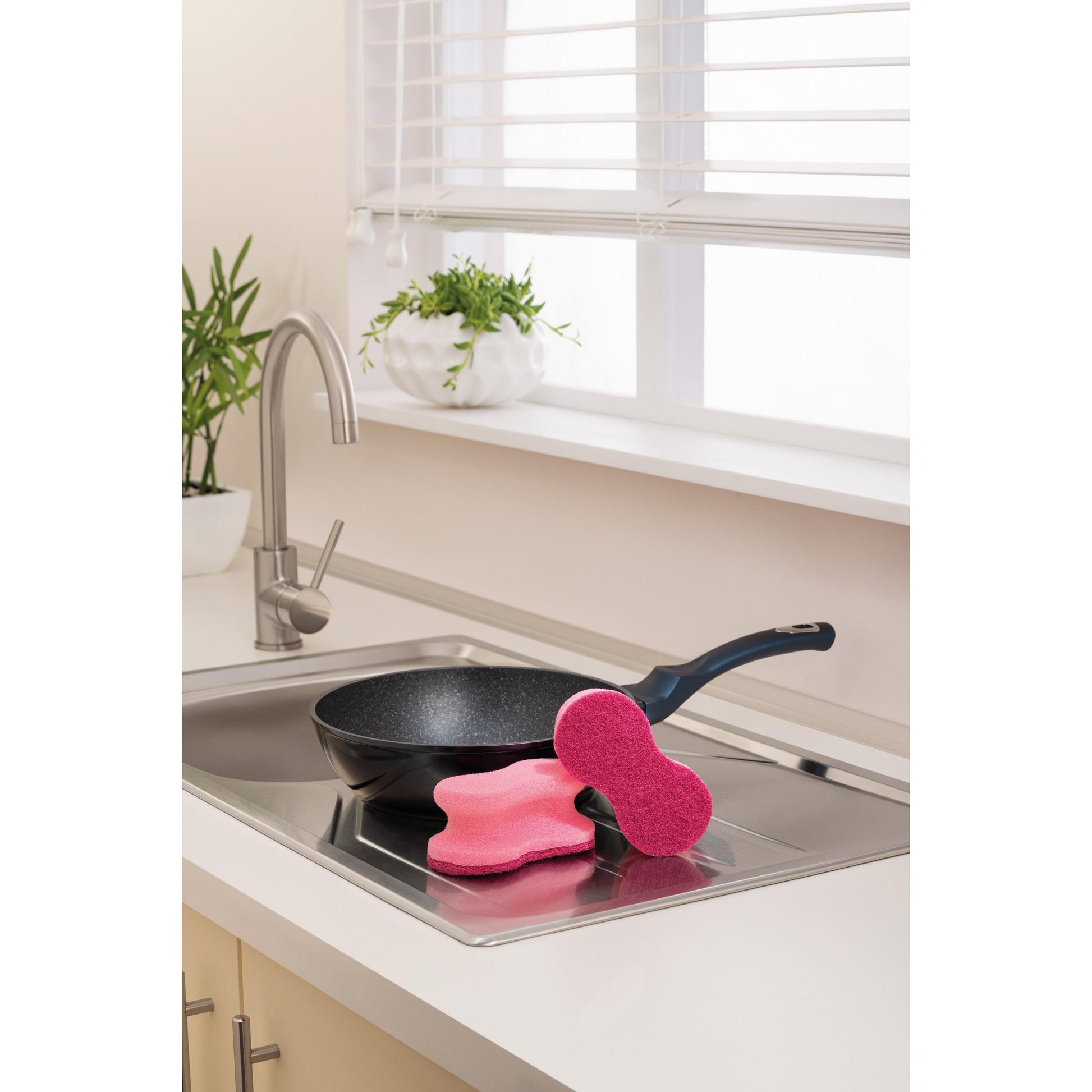 Eine schwarze Bratpfanne liegt in einer Küchenspüle, auf deren Rand zwei rosafarbene Scotch-Brite® Delicate Color Comfort nicht kratzender Reinigungsschwamm (2 Stück, 3M Deutschland GmbH) liegen; die weiße Arbeitsplatte und Topfpflanzen in der Nähe vervollständigen die Szene.