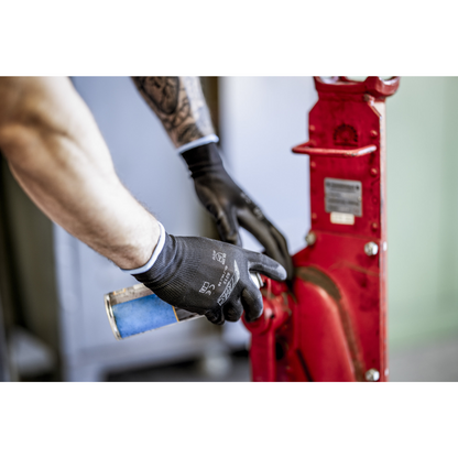 Een persoon met NITRAS nylonhandschoenen, zwart, PU-gecoat van AS Arbeitsschutz GmbH reinigt of smeert met een blauwe spuitbus een rood metalen machinedeel. Alleen armen en handen zijn te zien.