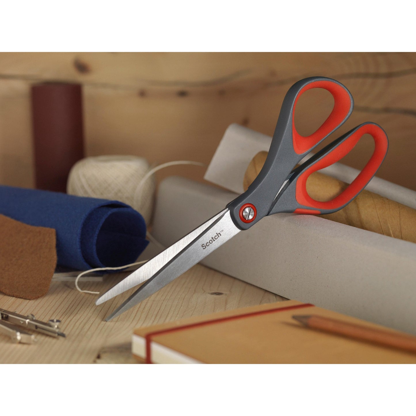 Die Scotch™ Präzisionsschere rot von 3M Deutschland GmbH mit grauem und rotem Griff liegt auf einer Holzfläche zwischen Stoffrollen, einer Spule mit weißem Garn, einem braunen Notizbuch und einem Bleistift.