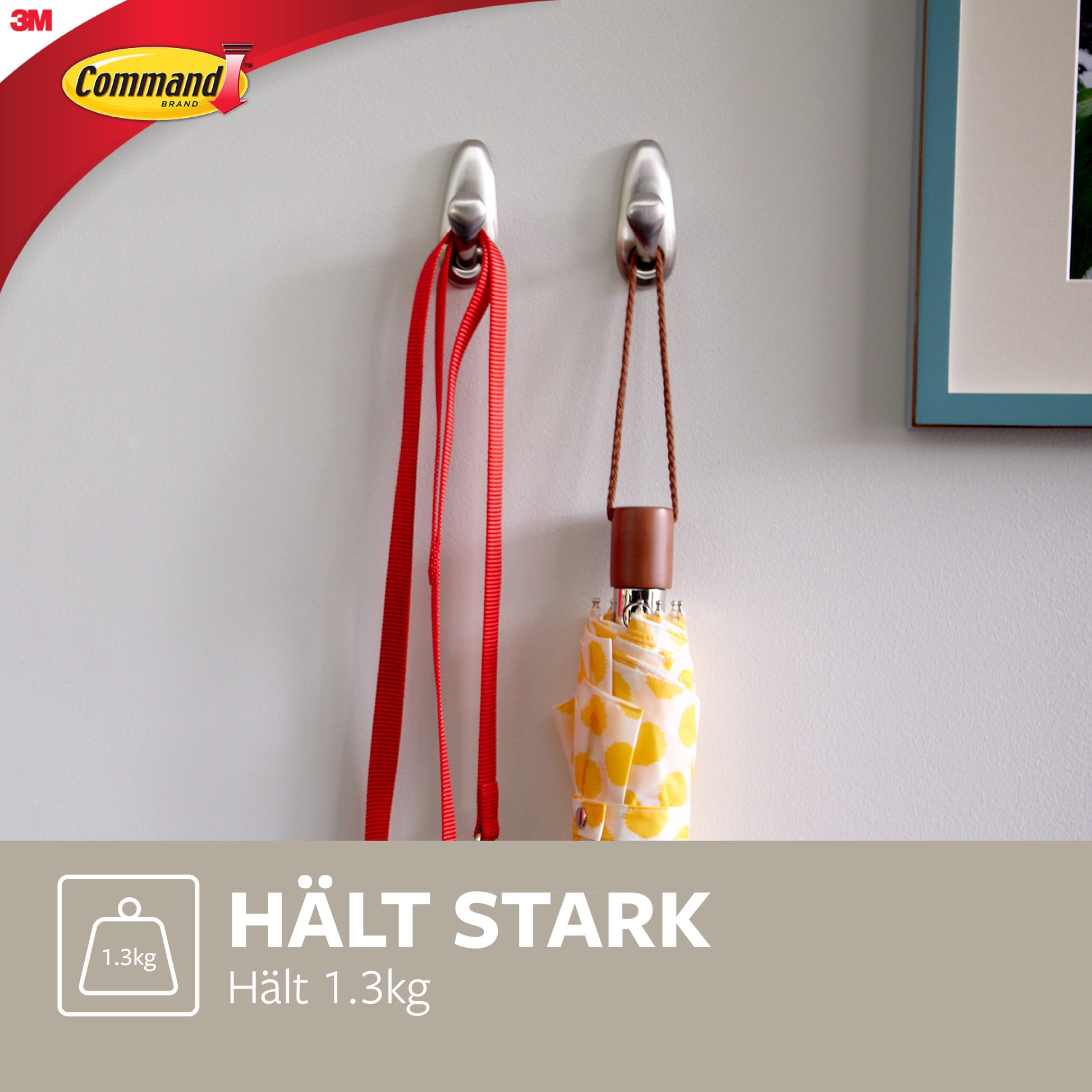 Zwei Command™ Metallhaken "klassisch" von 3M Deutschland GmbH sind an einer weißen Wand montiert und halten eine rote Leine und einen gelb gemusterten Regenschirm. Der Text lautet: "HÄLT STARK Hält 1.3kg" und zeigt das Command-Logo.