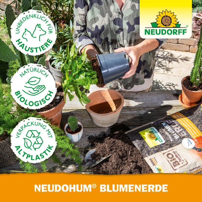 Een persoon bij het verpotten van een plant aan een tafel met aarde, gepotte tuinplanten, tuingereedschap, zakken potgrond en een NeudoHum BlumenErde-zak van Neudorff Shop. Naast het Neudorff-logo verschijnen de symbolen voor milieuvriendelijkheid en huisdiervriendelijkheid.