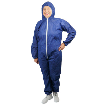Een persoon staat glimlachend in een ARNOMED COVER PP-OVERALL XTRA STRONG van ARNOWA GmbH, een blauwe, extra sterke wegwerpoverall met capuchon en witte ritssluiting aan de voorkant, gecombineerd met witte schoenen, voor een eenvoudige witte achtergrond.