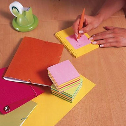 Iemand schrijft op een roze 3M Nederland B.V. Post-it® Notes kubus, Ultra-kleuren, aan een houten bureau met stapels van 76x76 mm plaknotities (450 vel/blok), een notitieblok, mappen en een groene plakbandhouder.