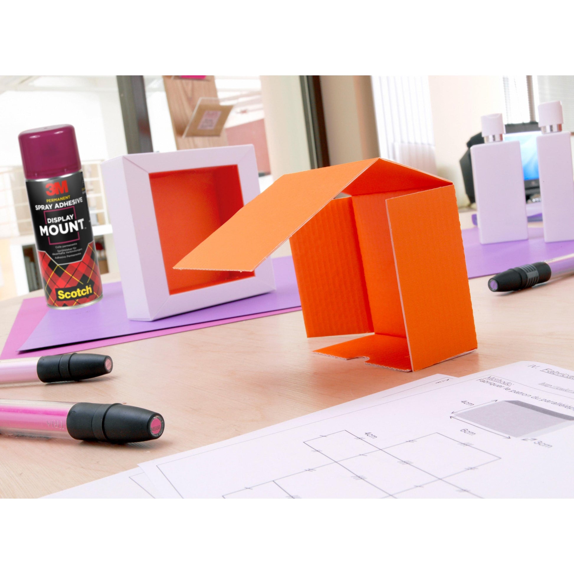Ein Tisch mit einem orangefarbenen Kartonmodell, Markern, 3M™ Scotch® DisplayMount™ Klebespray (400 ml, 1 Dose/Packung) von 3M Deutschland GmbH, weißen Schachteln und technischen Zeichnungen in einem hellen Innenarbeitsraum für den professionellen Einsatz.