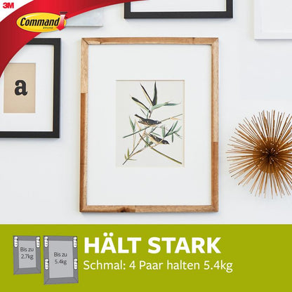 Een ingelijste botanische print hangt aan een witte muur met decor, waaronder goud en ingelijste letters. Tekst: "HOUDT STERK, Smal: 4 paar houden 5,4kg." Smalle Command™ Bevestigingsstrips voor schilderijen (M) 17207 van 3M Deutschland GmbH bevestigen smalle lijsten veilig.