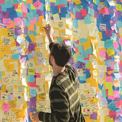 Een persoon in een gestreept overhemd staat bij een muur met handgeschreven berichten en tekeningen en grijpt naar een 3M Nederland B.V. Post-it® Notes kubus in Ultra-kleuren (76x76 mm, 450 vel, 100% PEFC), om deze tussen de kleurrijke blaadjes te plaatsen.