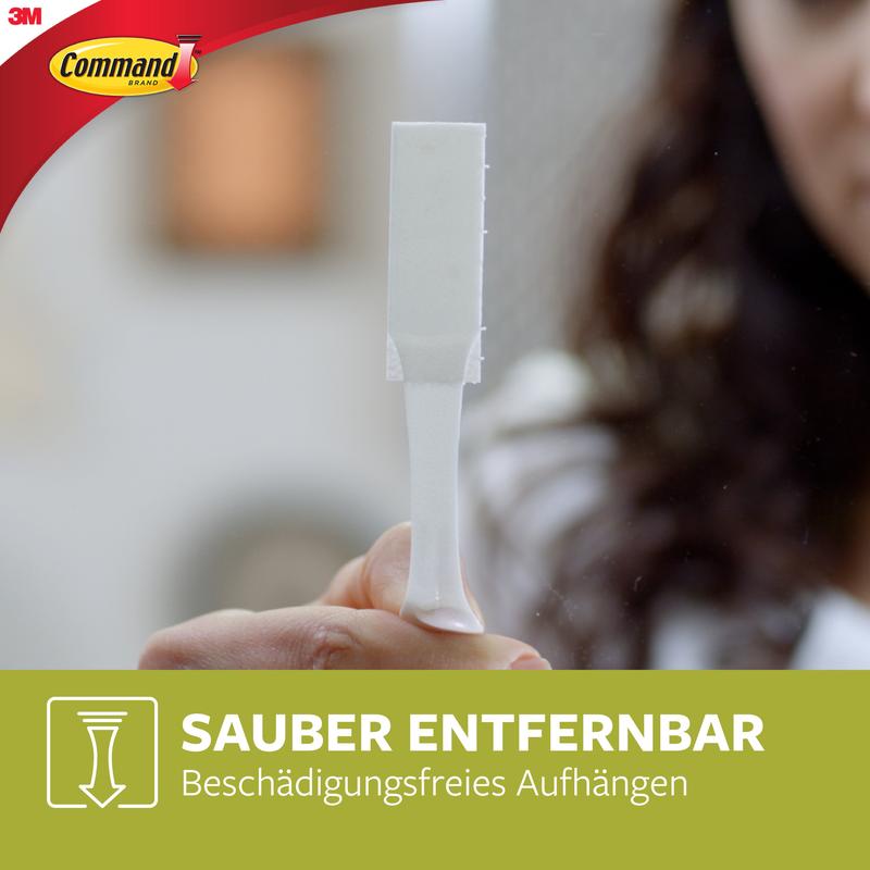 Een persoon houdt een witte Smalle Command™ Bevestigingsstrip (M) 17207 van 3M Deutschland GmbH rechtop. Het Command-logo en de Duitse tekst over smalle schilderijmontage en residuvrije verwijdering zijn te zien voor een wazige achtergrond.
