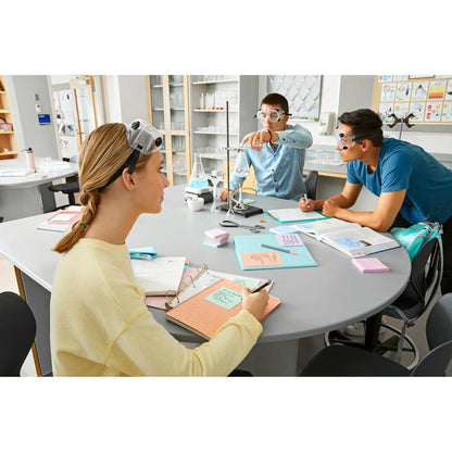 Drie studenten met veiligheidsbrillen zitten aan een laboratoriumtafel met leerboeken, laboratoriumapparatuur en 3M Deutschland GmbH Post-it® Super Sticky Notes (76 mm x 127 mm, 90 vel/Blok, 100% PEFC) en maken aantekeningen, terwijl één een klem afstelt en de anderen aandachtig toekijken.