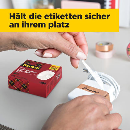 Een persoon wikkelt een wit etiket met zwarte letters om een witte oplader, terwijl er op de tafel een doos Scotch® Super-Hold Secure Tape, 1 rol, 19 mm x 25,4 m van 3M Deutschland GmbH staat. Op de achtergrond zijn wazige kantoormaterialen en een gele Duitse vlag te zien.