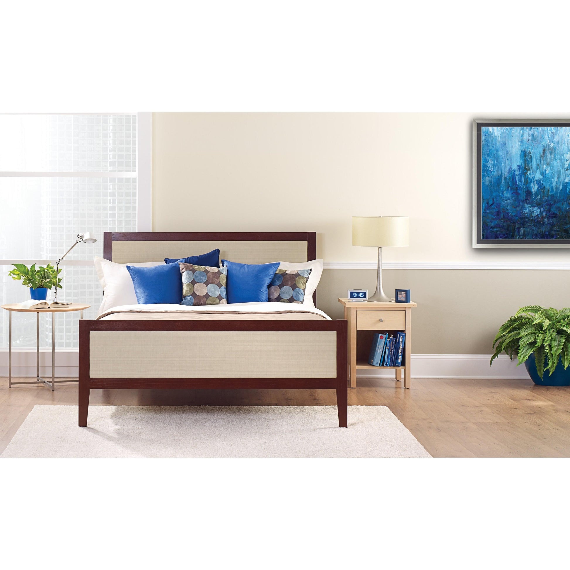 Ein ordentlich gemachtes Bett mit weißen und blauen Kissen steht vor einer hellen Wand, die mit ScotchBlue™ Malerabdeckband "Multi-Surface" der 3M Deutschland GmbH beklebt ist. Auf einem Nachttisch stehen eine Lampe, eine Uhr, Bücher und eine Topfpflanze neben dem hellen Fenster.