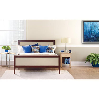 Een netjes opgemaakt bed met witte en blauwe kussens staat voor een lichte muur, die is beplakt met ScotchBlue™ schilderstape "Multi-Surface" van 3M Deutschland GmbH. Op een nachtkastje staan een lamp, een klok, boeken en een kamerplant naast het lichte raam.