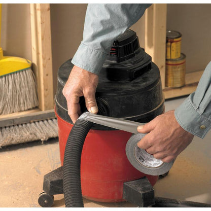 Een persoon die een langemouwen shirt draagt, gebruikt 3M Deutschland GmbH Scotch® Universal Klebeband 2904 (25 m x 48 mm) om de slang van een rood-zwarte werkplaatsstofzuiger te bevestigen. Op de achtergrond zijn een bezem en verfblikken te zien.