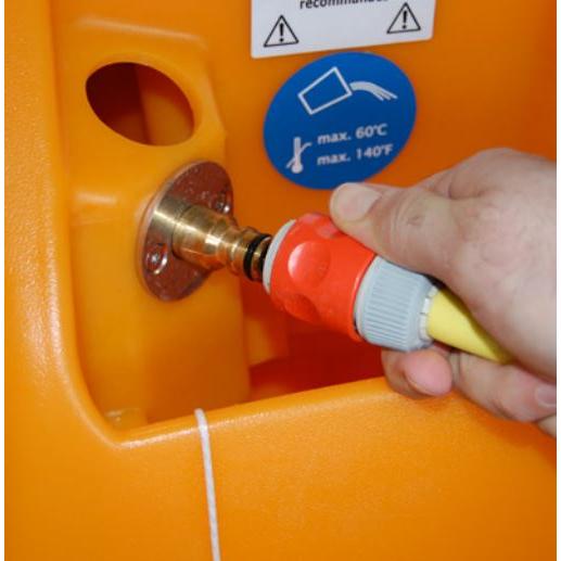Een hand verbindt een slang met een rood en grijs aansluitstuk met een metalen fitting aan een oranje TASKI swingo 2100 ?icro AquaStop van Diversey Deutschland GmbH & Co. OHG. Een blauwe sticker in de buurt toont een maximale temperatuur van 60°C/140°F aan.