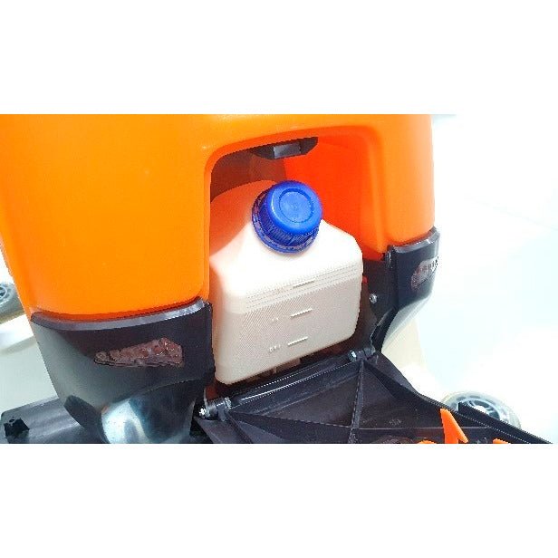 Een close-up van de Ultimaxx Intellidose+ kan van Diversey Deutschland GmbH & Co. OHG toont de witte vierkante container met blauwe schroefdop in een oranje vak, omringd door zwarte en metalen onderdelen.