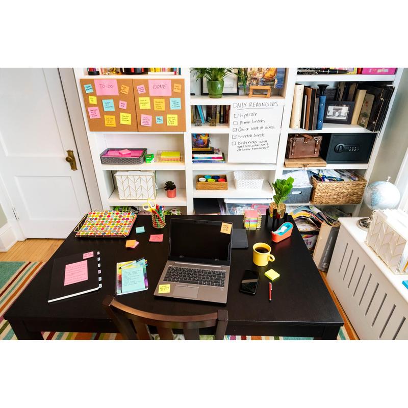 Een bureau met laptop, notitieboeken, telefoon, pennen, muis, gele beker en Post-it® Super Sticky Z-Notes (Boost Collection) van 3M Deutschland GmbH. Daarachter bevinden zich planken met boeken, opbergdozen en een whiteboard met geheugensteuntjes en kleurrijke kleefnotities.