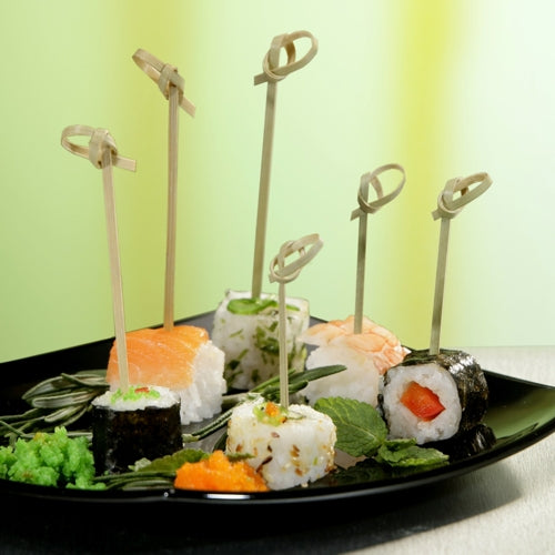 Een zwart bord met verschillende sushi, elk stuk doorboord met een PAPSTAR 40 Fingerfood-prikker „pure“ van PAPSTAR GmbH, gegarneerd met kruiden en kuit, op groen-gele ondergrond – ideaal om cateringaccessoires in de schijnwerpers te zetten.