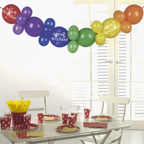 Een witte tafel met rood gestippeld feestservies, gele bestekken en bekers wordt bekroond door de PAPSTAR ballonkrans 2 m "Happy Birthday" van PAPSTAR GmbH - een feestelijke verjaardagsfeestdecoratie die in het zonlicht door witte luiken straalt.