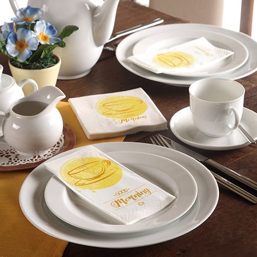 Een keurig gedekte tafel met wit servies, een theepot, blauwe bloemen en 3-laags PAPSTAR-servetten (33 x 33 cm) van de PAPSTAR GmbH met een gele koffiekop en het opschrift „Morning“.