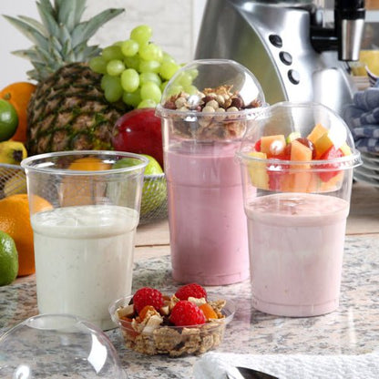 Drie smoothie-bekers staan op een toonbank, twee met fruit en granola in koepeldeksels. De deksels zijn gemaakt van Starpak 50 inzetten, PET "Hurricane" rond Ø 9 cm, glashelder. Op de achtergrond staan verse ananas, druiven en een blender.