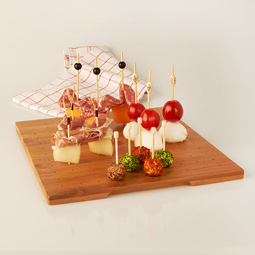 Der PAPSTAR Halter für Fingerfood-Picker aus Bambus „pure“ (25x30 cm, Schrumpffolie, 1 Stück) von PAPSTAR GmbH präsentiert Appetizer-Spieße mit Tomaten, Mozzarella, Schinken, Melone, Käsekugeln und Oliven; im Hintergrund ein rot-weißes Tuch.
